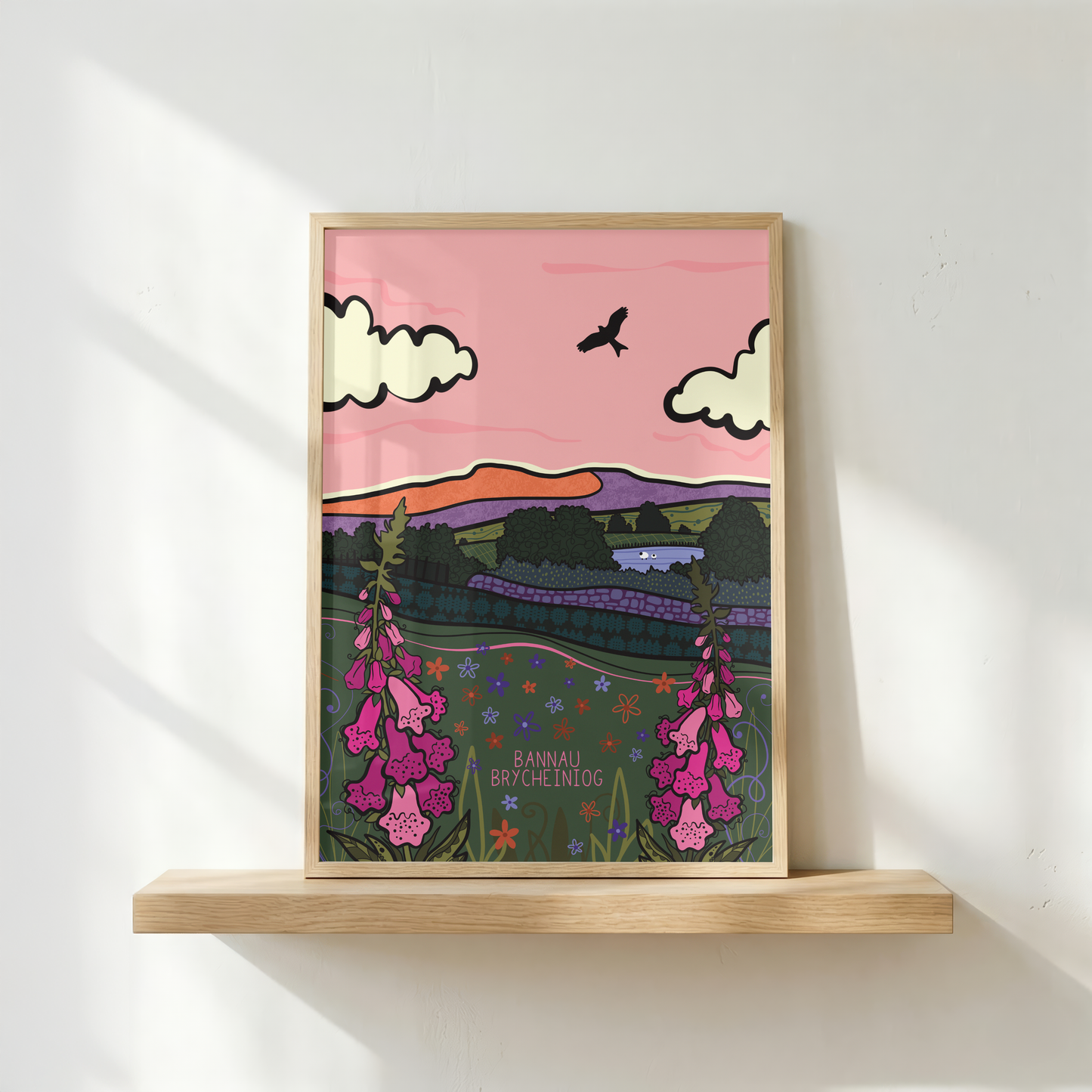 A framed art print featuring a stylized landscape illustration with pink sky, white clouds, a bird, purple hills, and pink foxglove flowers, displayed on a light wood floating shelf against a white wall with soft window shadows.