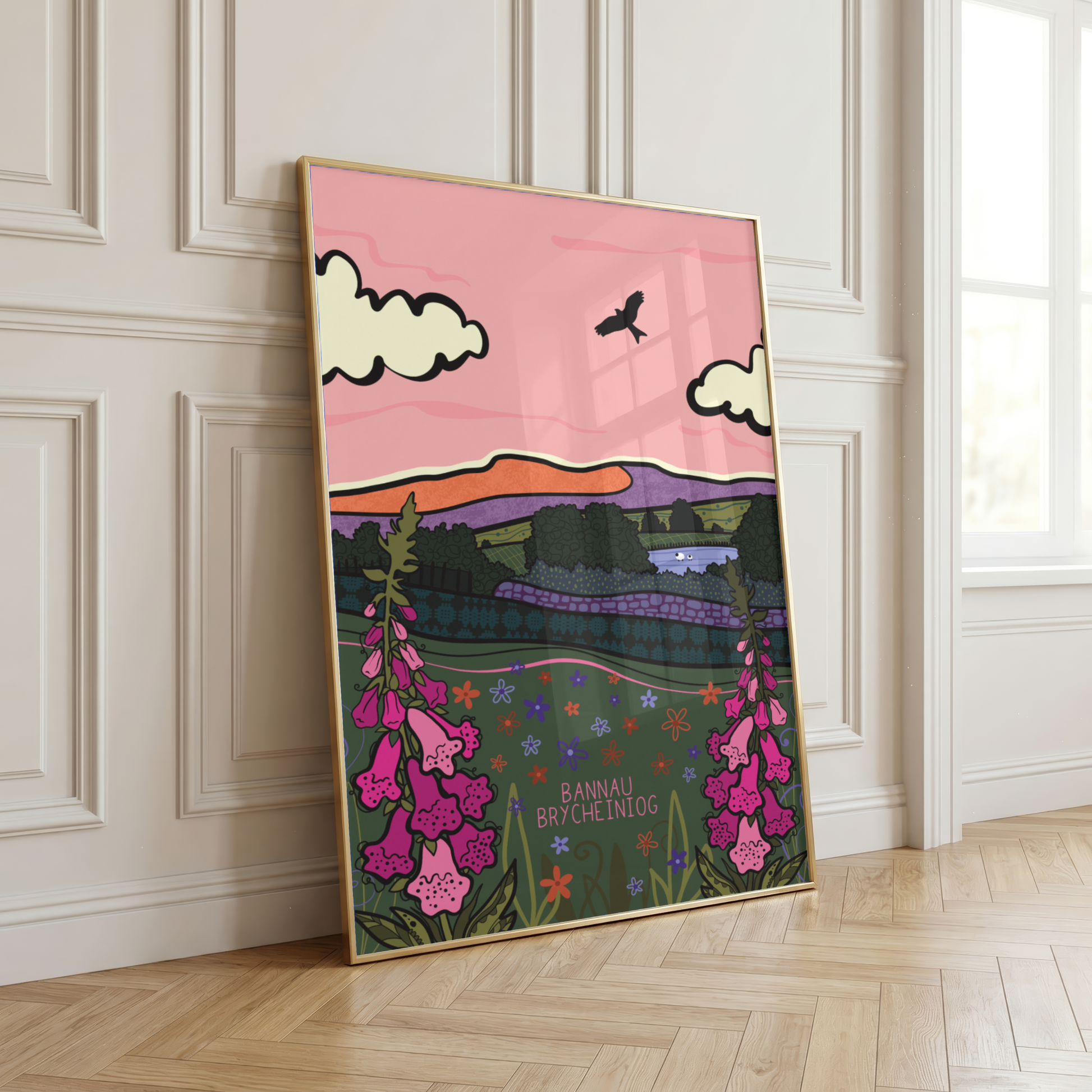 A framed art print leaning against a white paneled wall on a light wood floor. The print features a colorful landscape with pink sky, white clouds, a bird, green hills, and pink flowers, with the text 'BANNAU BRYCHEINIIG' in the centRE